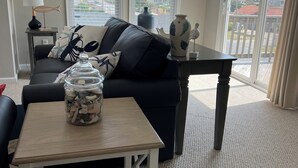 A living room with a black sofa, a coffee table with a jar of seashells, a lamp, and a view of the neighborhood through large windows. - SPACIOUS FAMILY FRIENDLY HOME, 300 STEPS TO BEACH, PRIVATE POOL/HOT TUB (Kill Devil Hills)