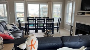 A modern living room with a high ceiling, a large window, a fireplace, and a dining area. - SPACIOUS FAMILY FRIENDLY HOME, 300 STEPS TO BEACH, PRIVATE POOL/HOT TUB (Kill Devil Hills)