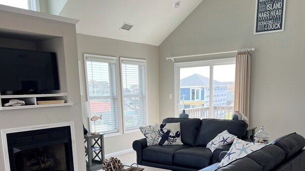 A modern living room with a large dark sofa, a fireplace, a flat-screen TV, and large windows offering a view of the city. - SPACIOUS FAMILY FRIENDLY HOME, 300 STEPS TO BEACH, PRIVATE POOL/HOT TUB (Kill Devil Hills)