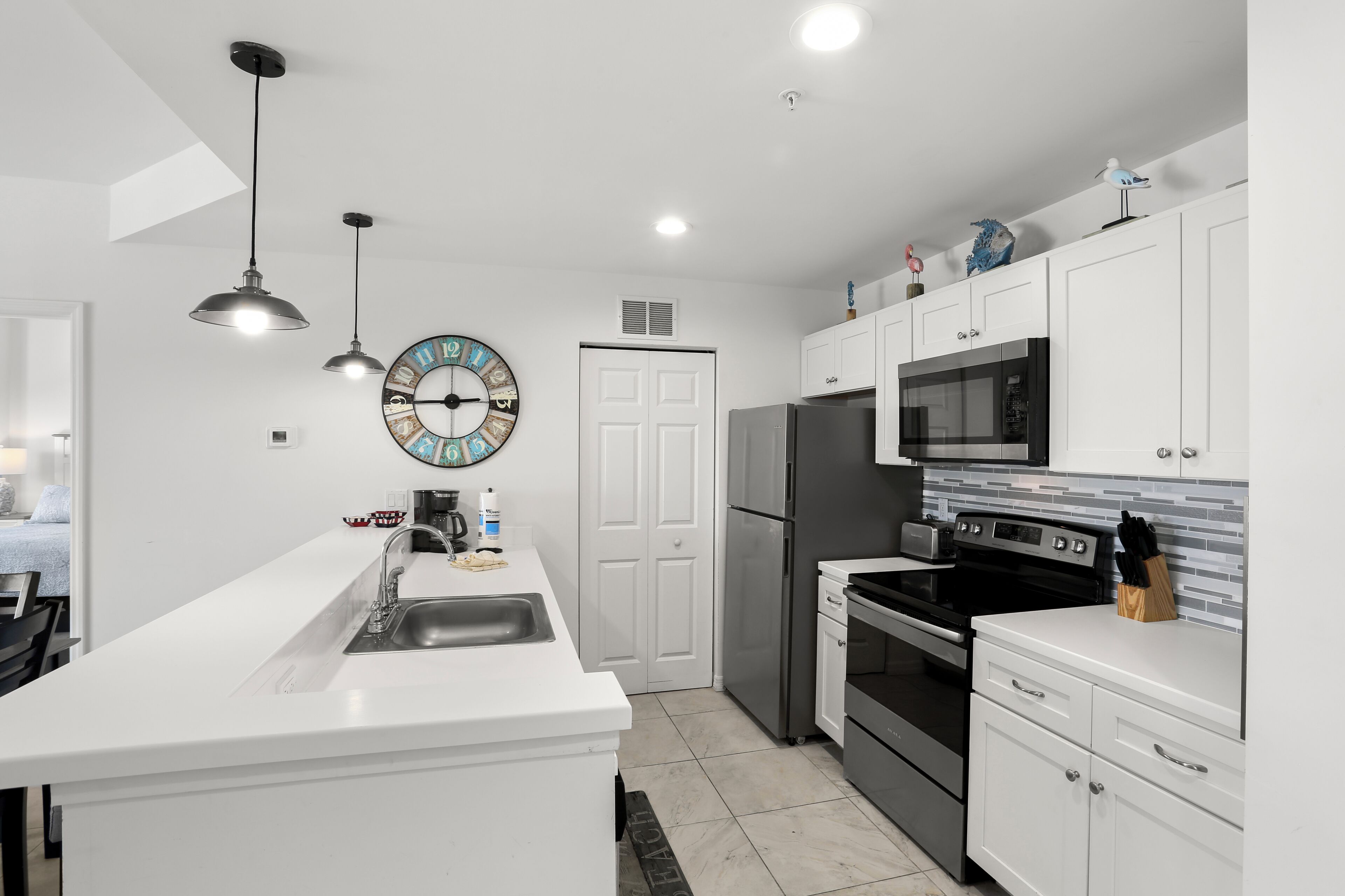 Uma cozinha moderna com armários brancos, eletrodomésticos de aço inoxidável, iluminação pendente e um relógio decorativo.