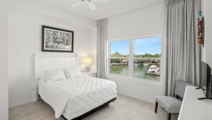 A bedroom with a bed, a desk, a chair, a TV, a ceiling fan, a window with a view of a marina, and a framed picture on the wall.