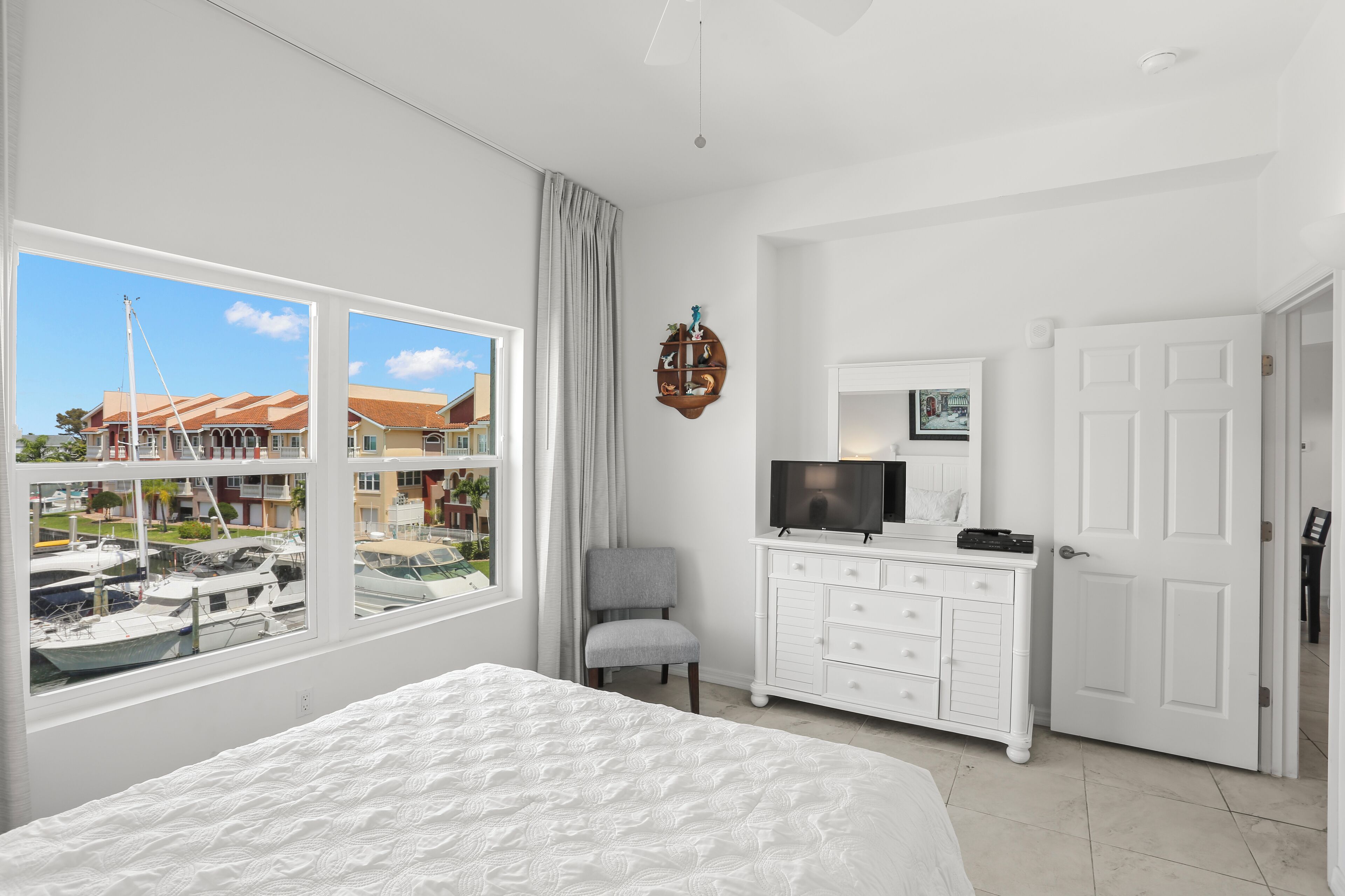 A bedroom with a large bed, a dresser, a television, and a view of boats through a window.