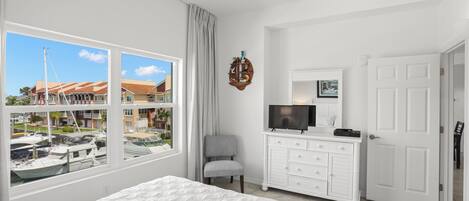 A bedroom with a large bed, a dresser, a television, and a view of boats through a window.