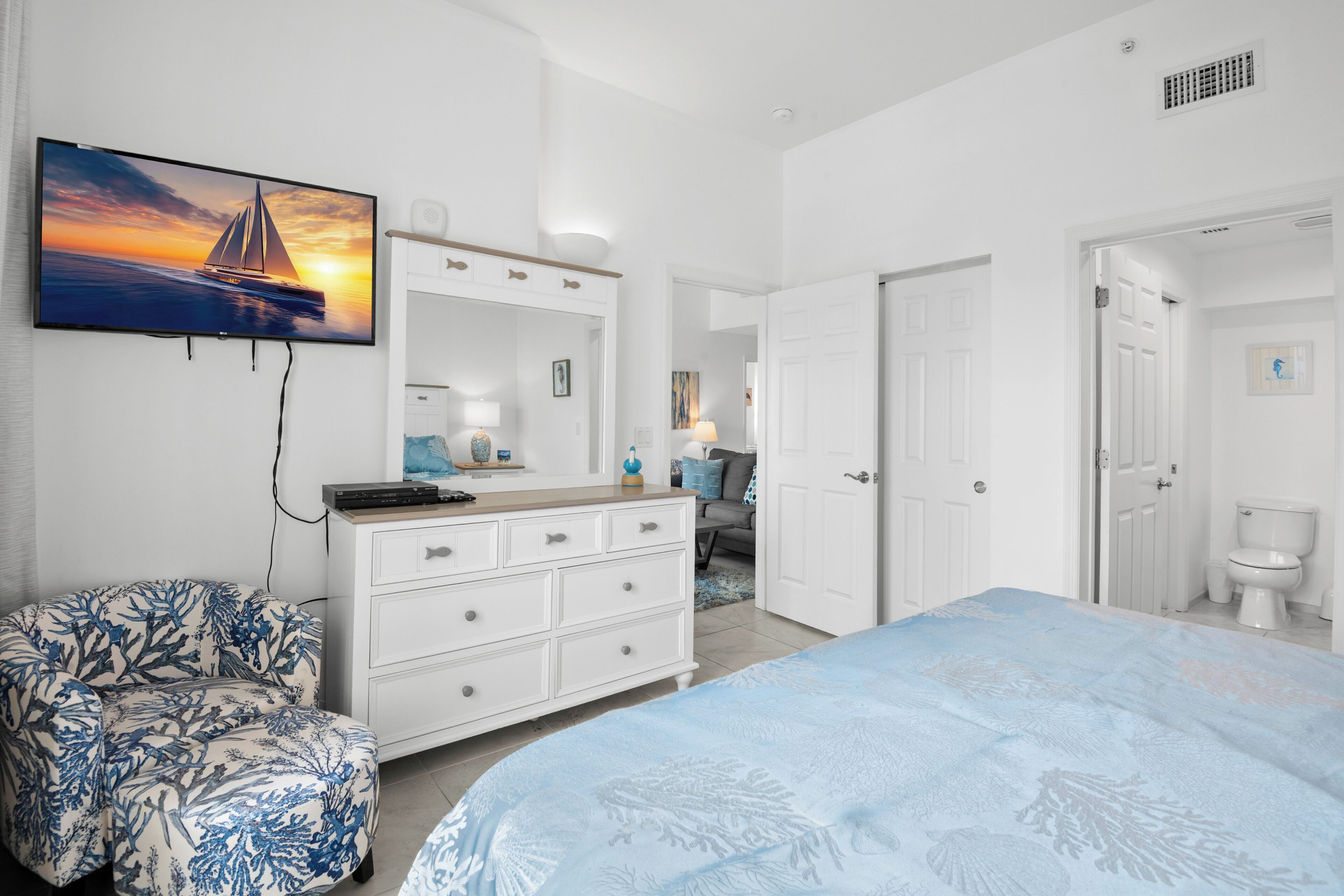 A bedroom with a bed, a dresser, a chair, a TV on the wall, and a door leading to another room.