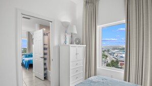 A bedroom with a bed, a dresser, a lamp, and a view of a cityscape through a window. - 🐾 Madeira Bay Resort I #1601 – Spacious, Pet-Friendly Escape by the Shore (Madeira Beach)