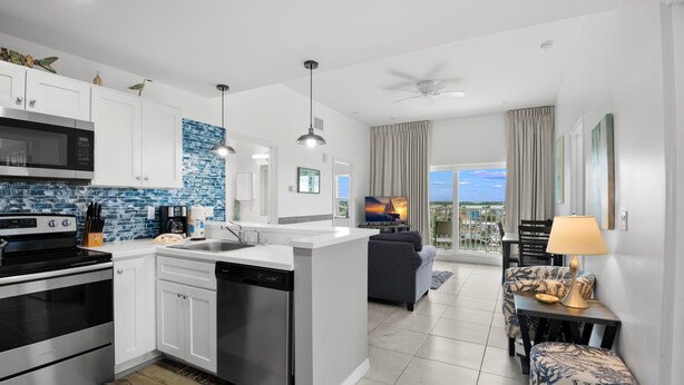 A modern kitchen with white cabinets, a built-in microwave, and a dishwasher. There is a dining area with a sofa and a table, and a balcony with a view. - 🐾 Madeira Bay Resort I #1601 – Spacious, Pet-Friendly Escape by the Shore (Madeira Beach)