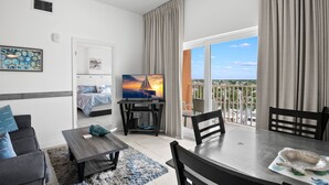 A modern living room with a TV, a dining table, and a balcony with a view. - 🐾 Madeira Bay Resort I #1601 – Spacious, Pet-Friendly Escape by the Shore (Madeira Beach)