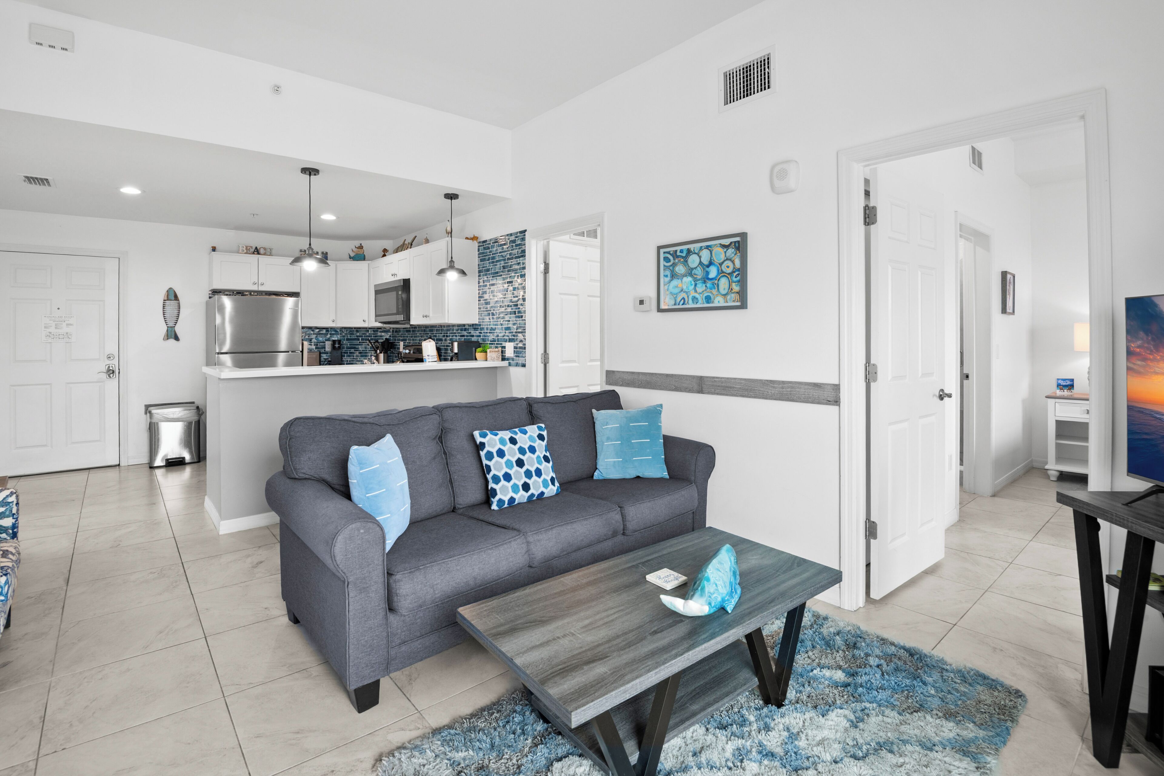 A modern living room with a grey sofa, blue patterned cushions, a wooden coffee table, and a kitchen in the background.