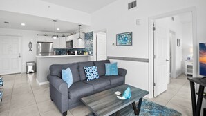 A modern living room with a grey sofa, blue patterned cushions, a wooden coffee table, and a kitchen in the background. - 🐾 Madeira Bay Resort I #1601 – Spacious, Pet-Friendly Escape by the Shore (Madeira Beach)