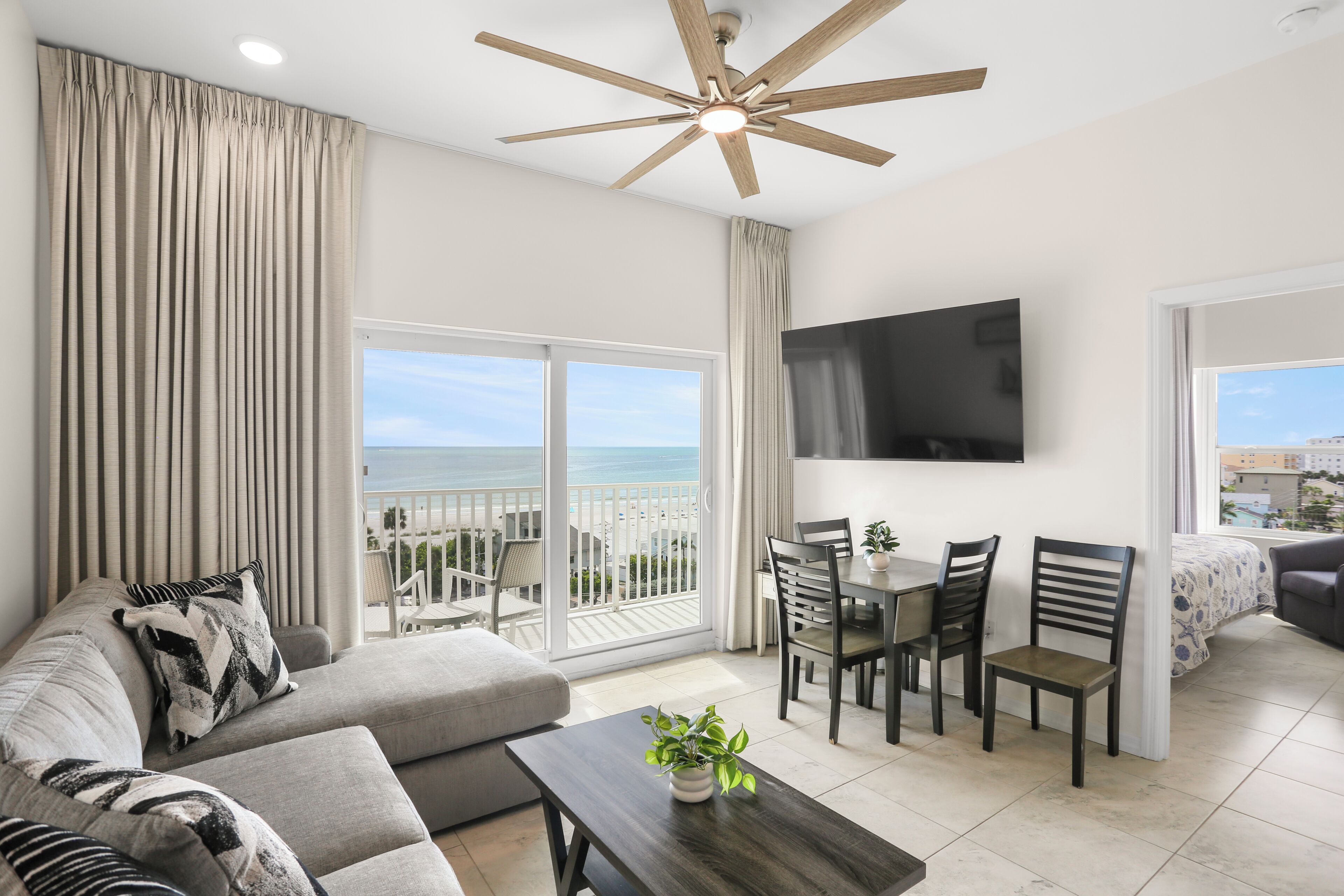 A modern living room with a sofa, dining table, chairs, a TV, and a balcony with a view of the ocean.