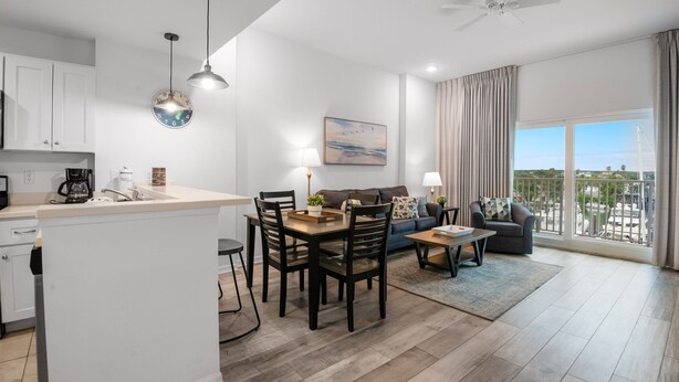 A modern apartment with a kitchen, dining area, and living room. It features a large window with a view, a ceiling fan, and a clock on the wall. - 🏝️ Madeira Bay Resort I #1403 – Cozy, Kid-Friendly (Madeira Beach)