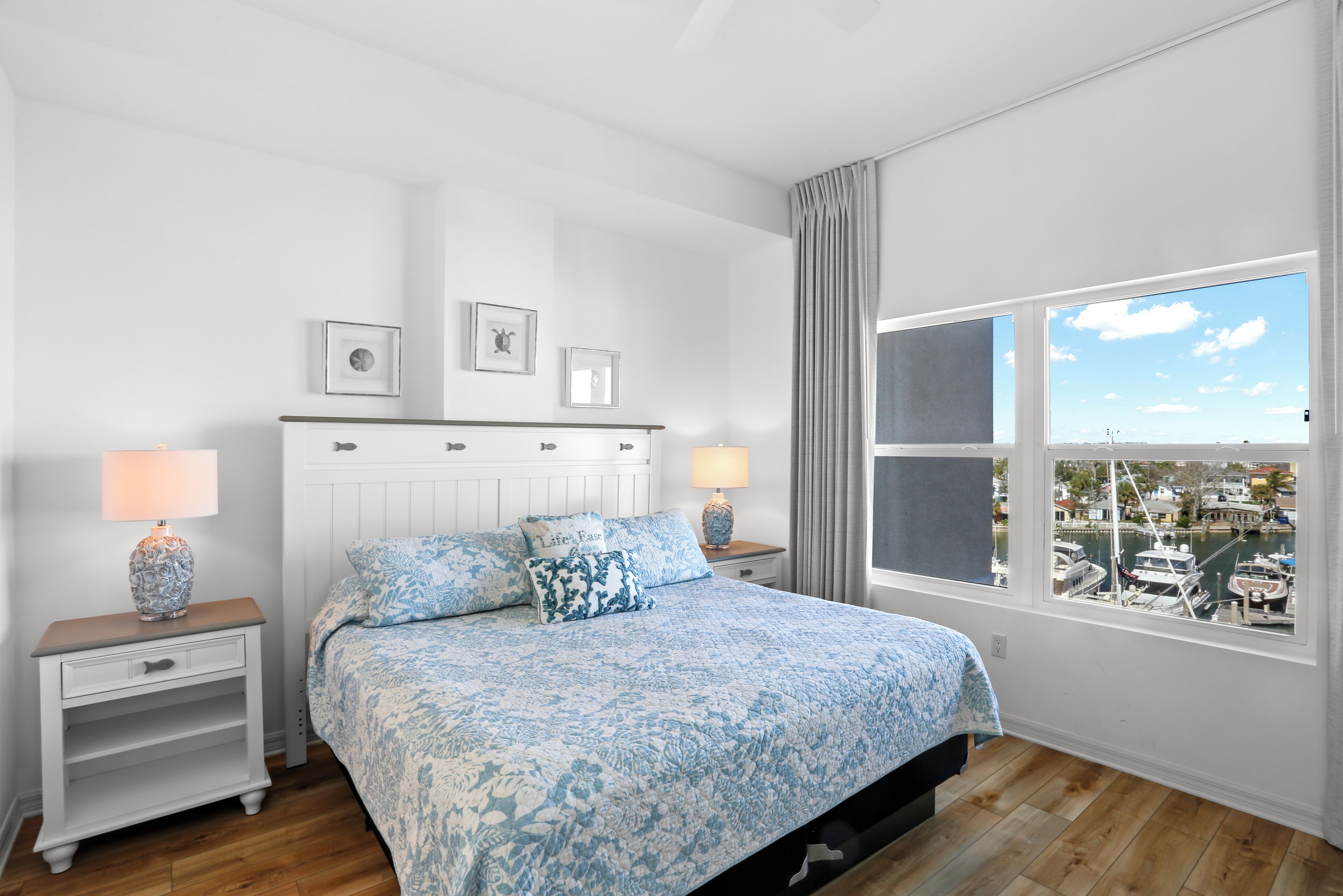 A bedroom with a large bed, two bedside tables, a window with a view of boats, and a white wall with three framed pictures.