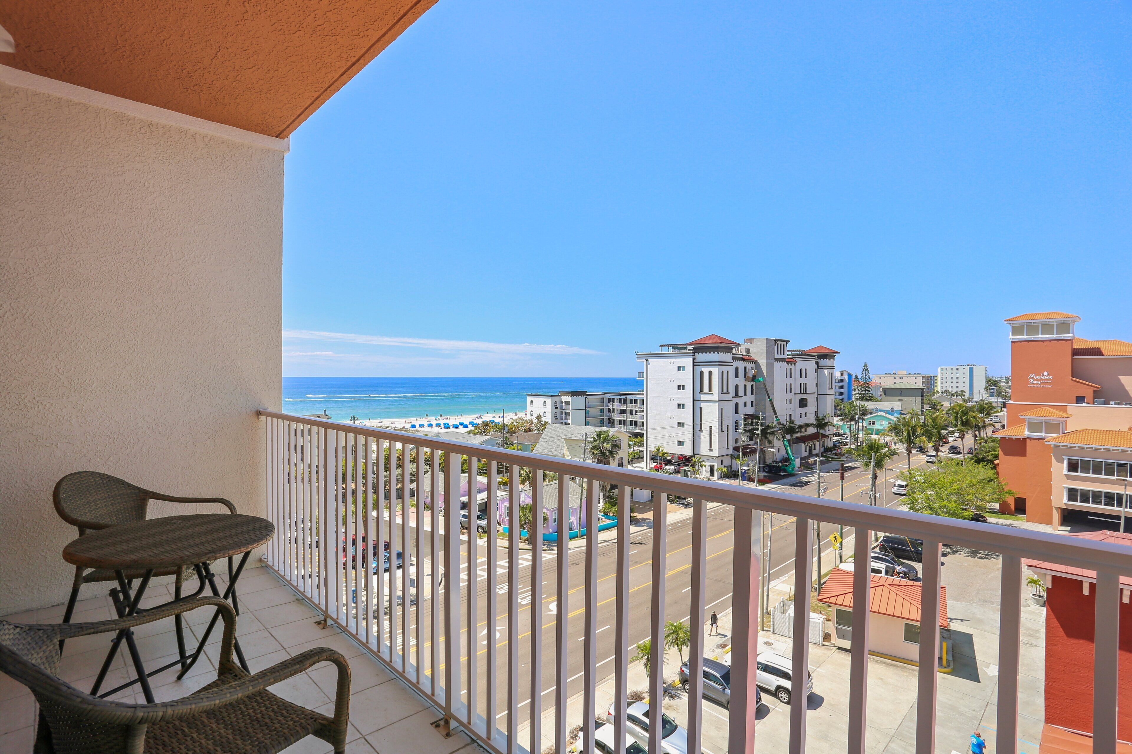 Un balcón con sillas de mimbre y una mesa, con vista a una playa y un paisaje urbano con edificios, bajo un cielo despejado.