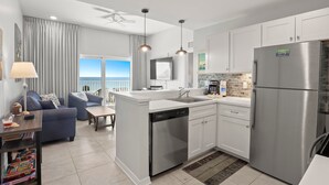 A modern kitchen with white cabinets, a stainless steel refrigerator, and a dishwasher. There is a dining area with a blue sofa and a table. - 🐾 Madeira Bay Resort I #1608 – Pet-Friendly, Professionally Sanitized & Steps from the Shore (Madeira Beach)