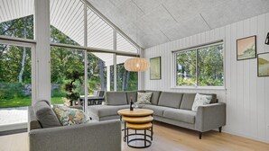 Ein modernes Wohnzimmer mit einem grauen Sofa, einem runden Holztisch und einer Hängeleuchte. Große Fenster bieten einen Blick ins Freie.