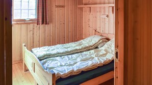 Un simple lit en bois avec un matelas vert, dans une pièce aux murs en bois et dotée d’une fenêtre ornée d’un rideau à carreaux.