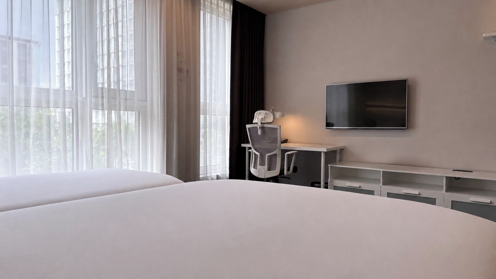 A hotel room with a large bed, a desk with a chair, a television mounted on the wall, and a window with sheer curtains.