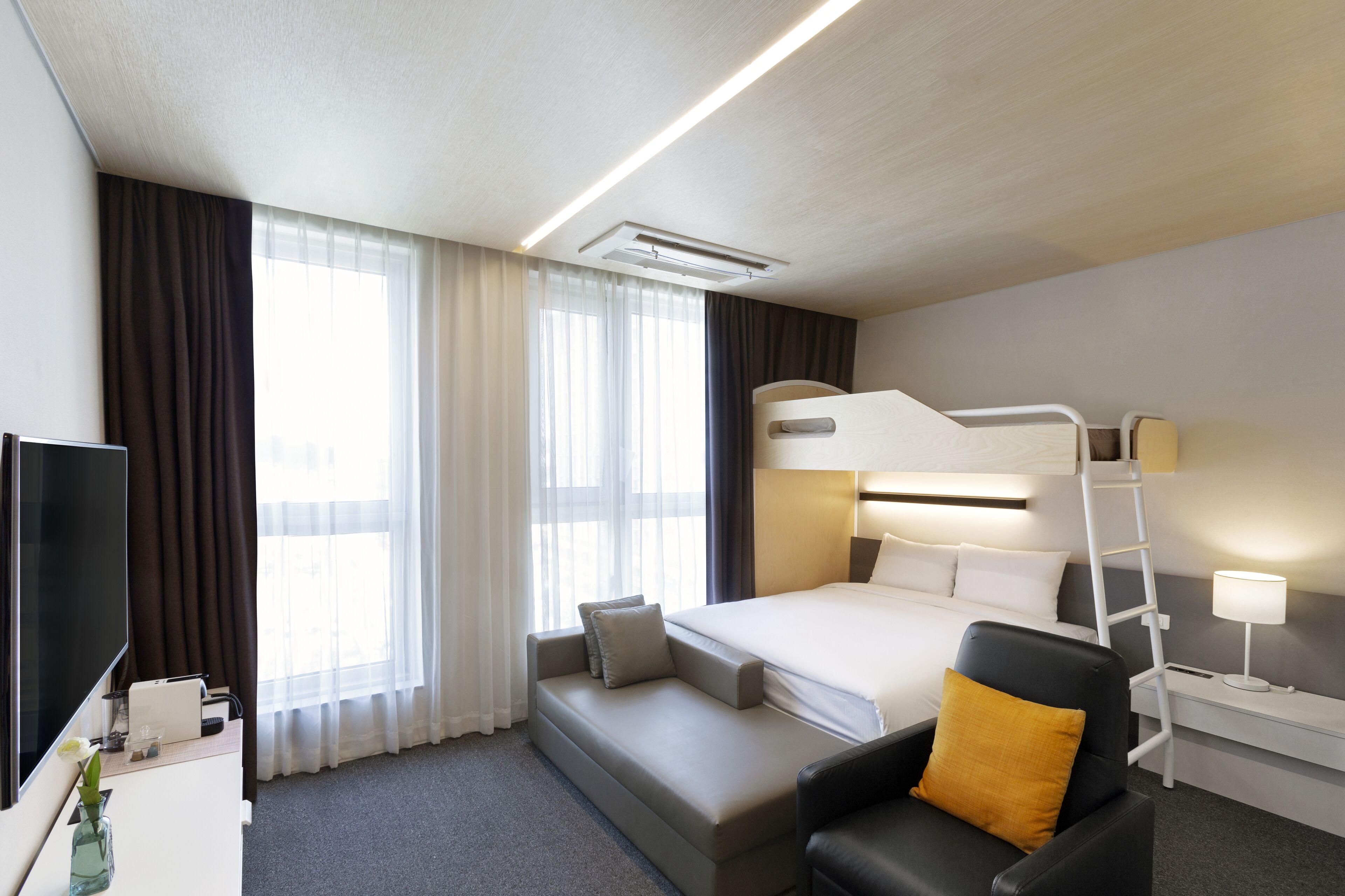 A hotel room with a bunk bed, a desk with a TV, a chair, a bedside table with a lamp, and a large window with curtains.