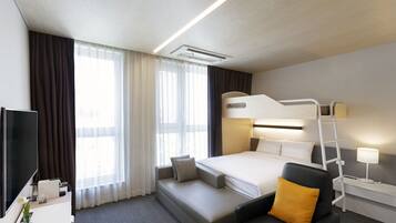 Habitación de hotel con literas, un escritorio con televisor, una silla, una mesita de noche con lámpara y un ventanal grande con cortinas.