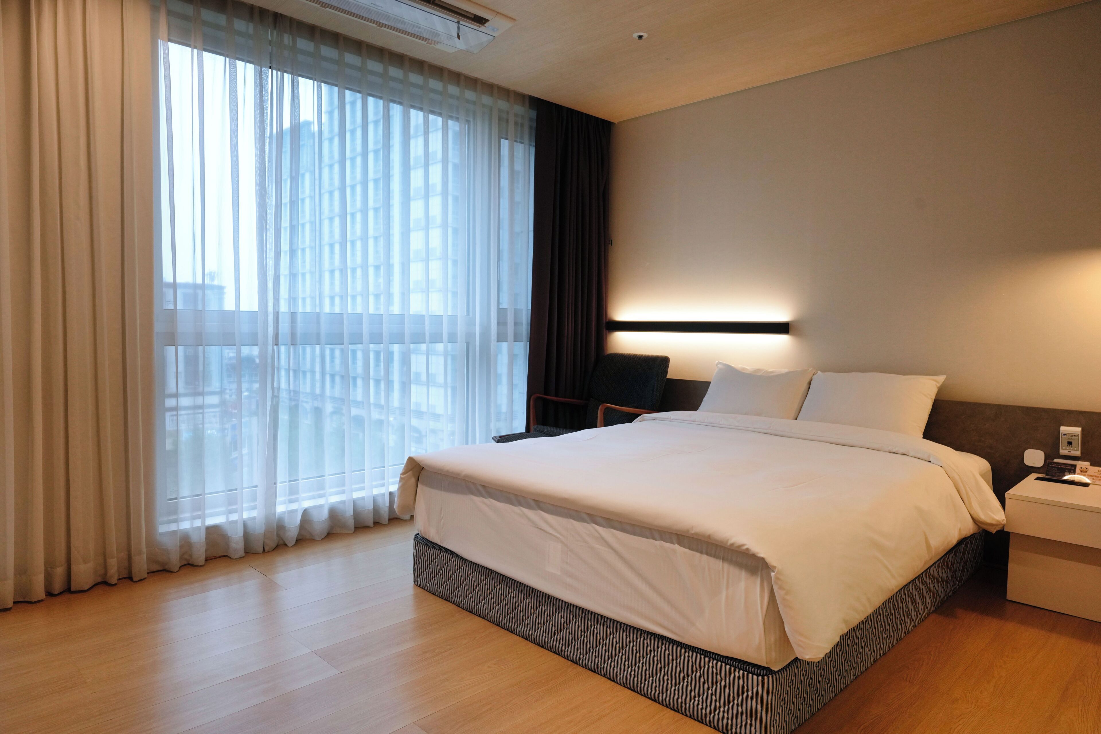 A modern hotel room with a large bed, a bedside table, a nightstand, and a window with sheer curtains.