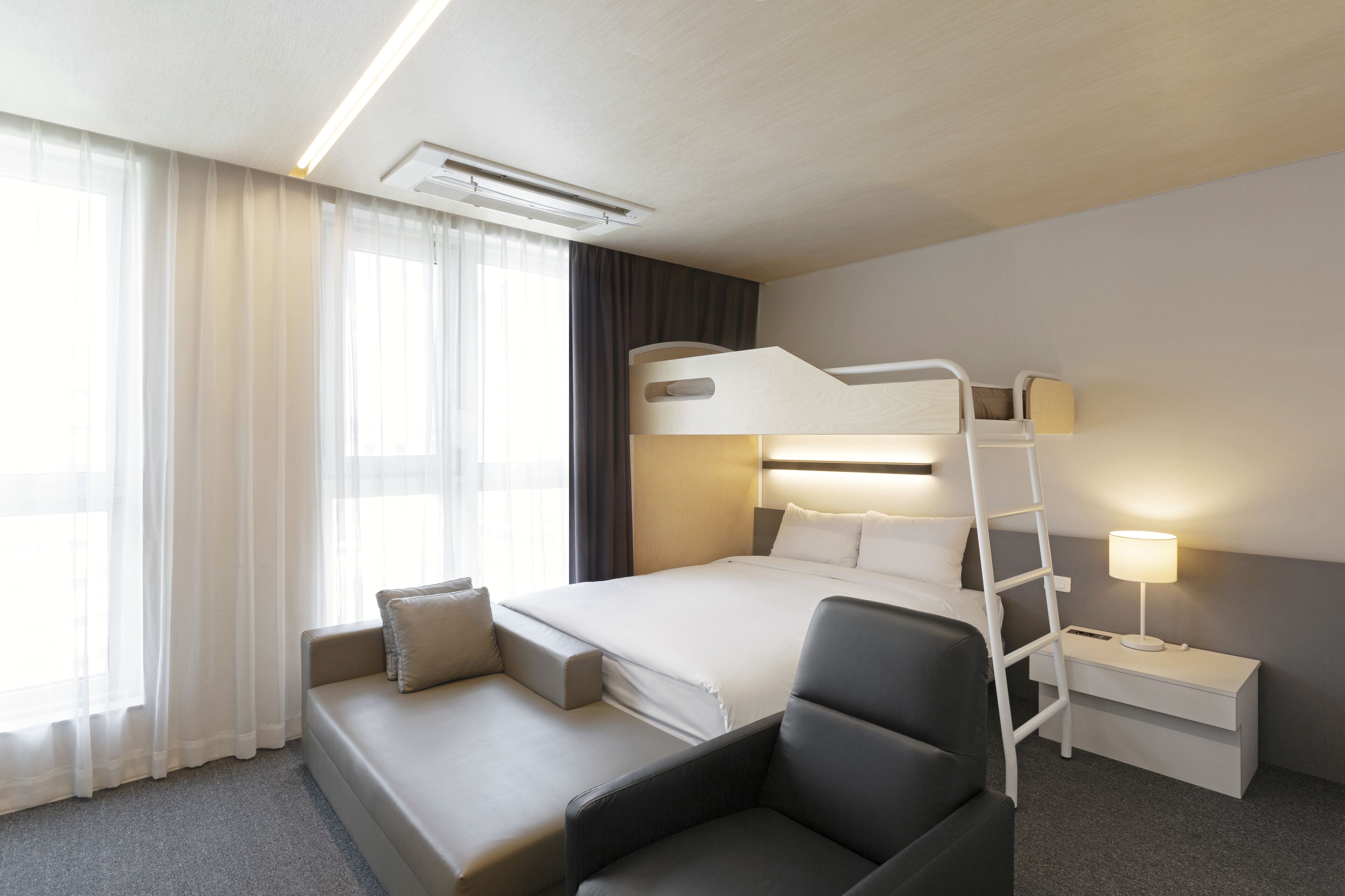 A modern hotel room with a bunk bed, a sofa, a desk with a lamp, and a large window with sheer curtains.