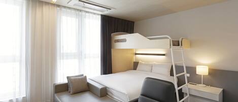 A modern hotel room with a bunk bed, a sofa, a desk with a lamp, and a large window with sheer curtains.