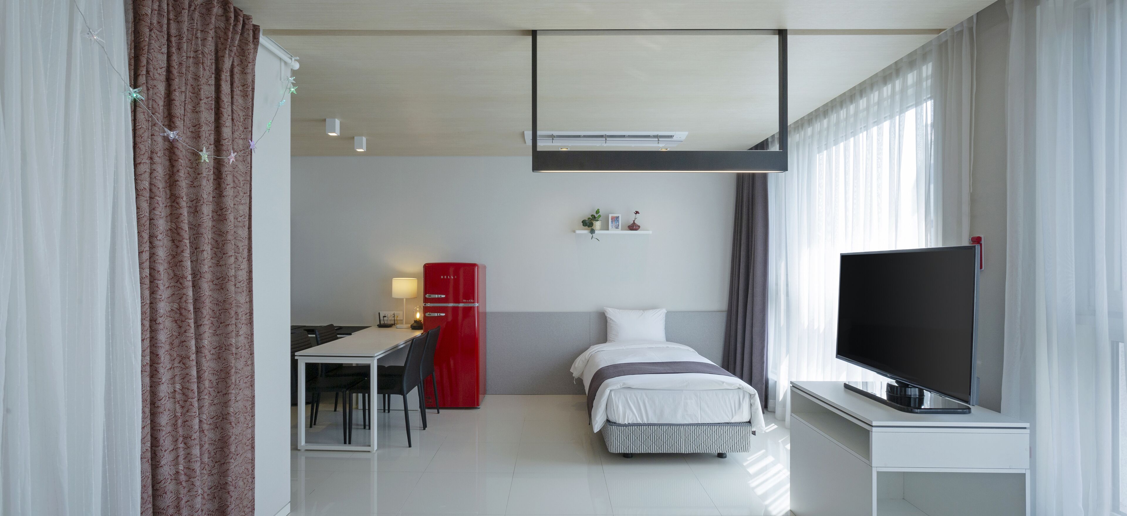 A modern hotel room with a bed, a desk, a red refrigerator, a TV, and a window with curtains.