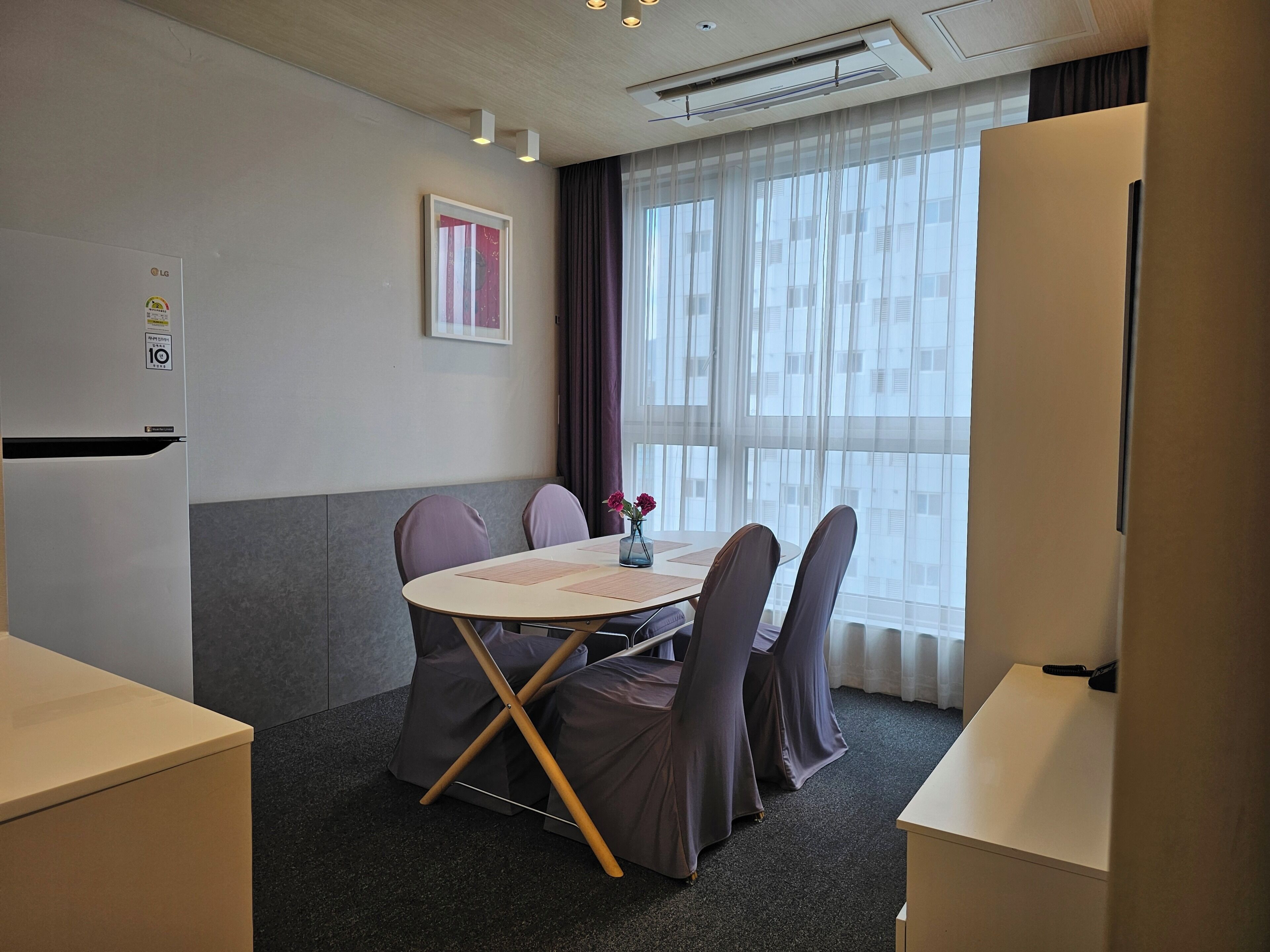 Un salón de conferencias con una mesa, sillas, un refrigerador y una ventana con cortinas.