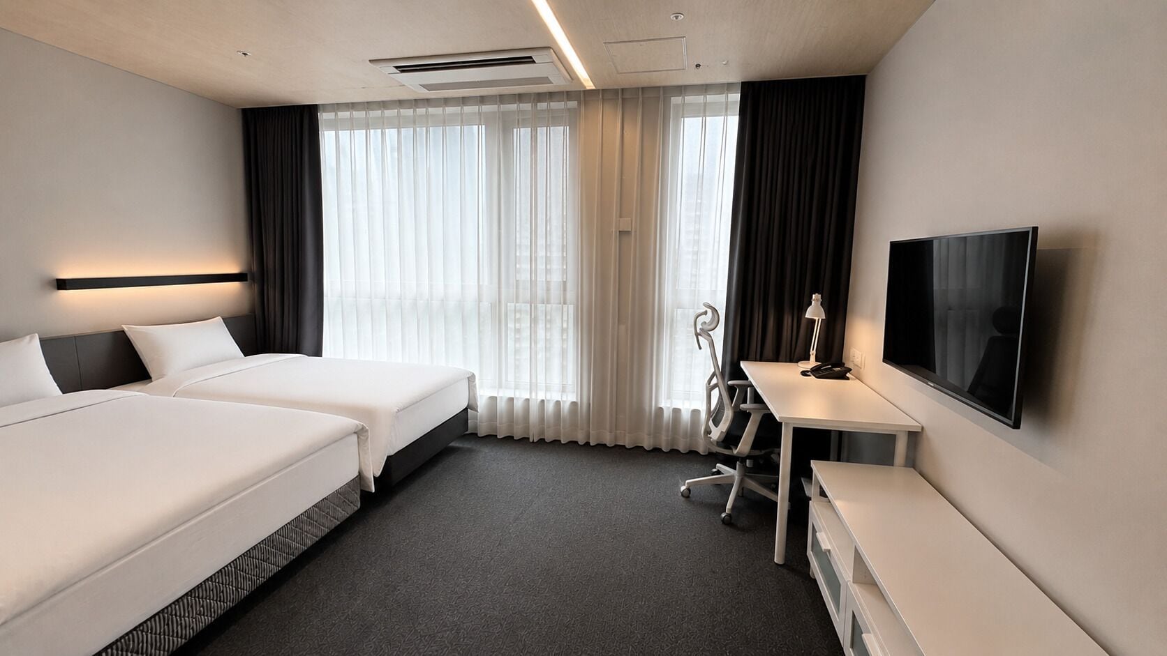 A hotel room with two beds, a desk, a chair, a television, and a large window with curtains.