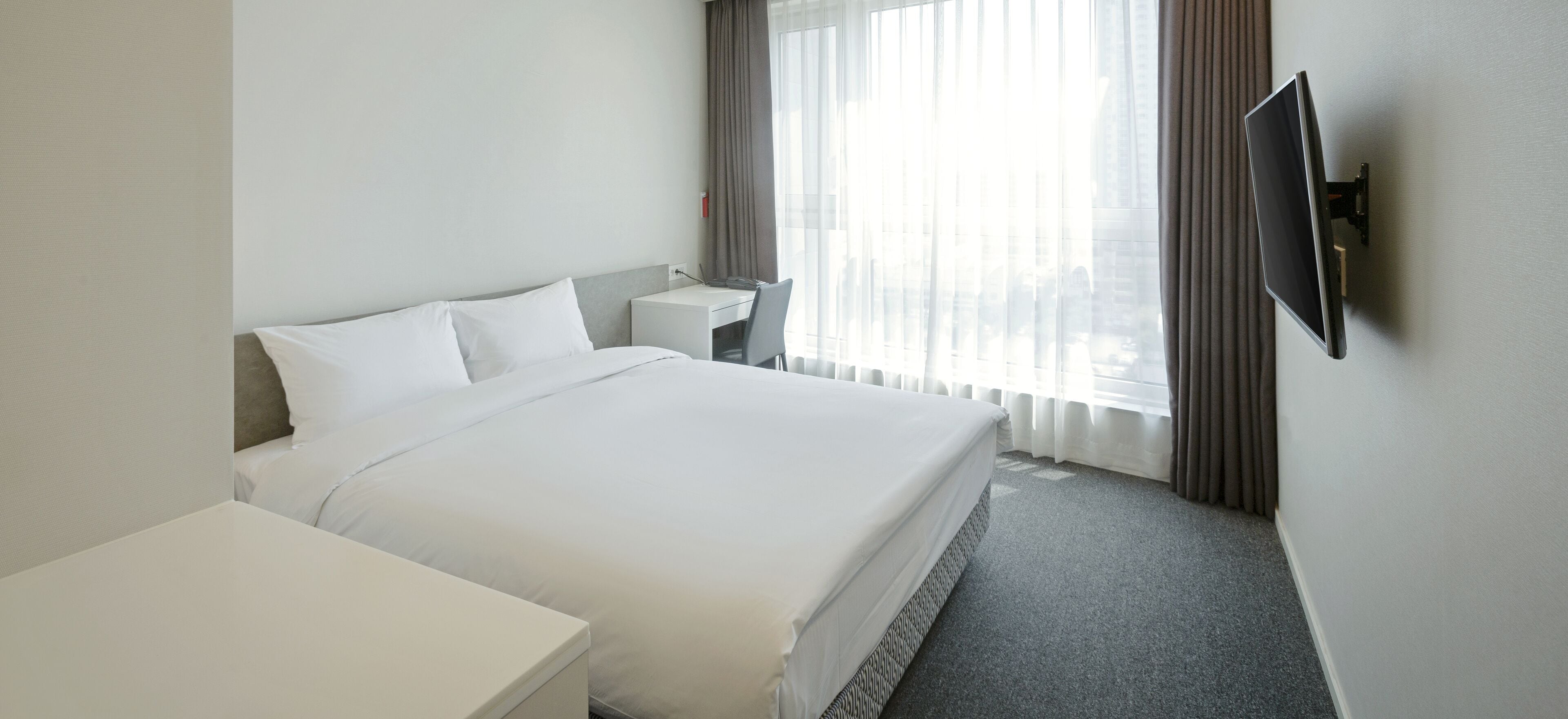 A hotel room with a large bed, a desk, a chair, a TV mounted on the wall, and a window with curtains.
