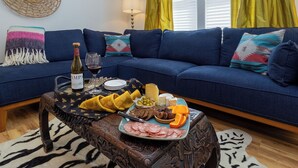 Una sala de estar acogedora con un sofá azul, una mesa de centro de madera con un plato de comida, una botella de vino y una lámpara.