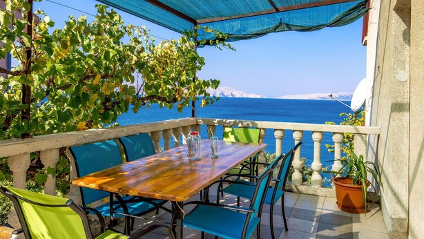 Een houten tafel met stoelen en een blauwe parasol op een balkon met uitzicht op zee.
