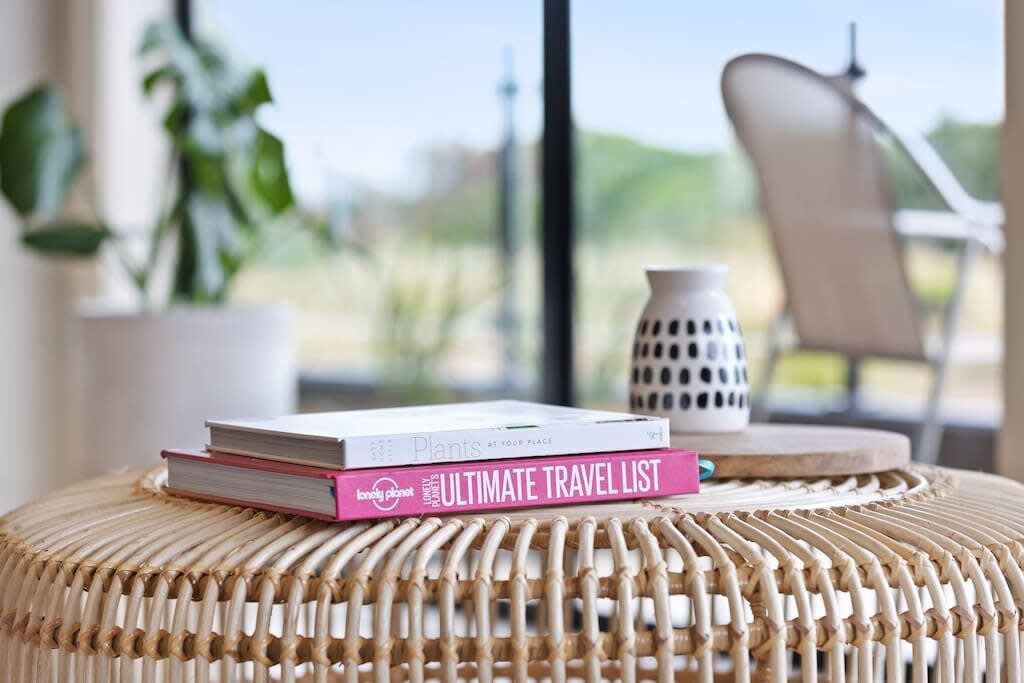 A wicker ottoman with books, a white vase, and a chair in a room with a view.