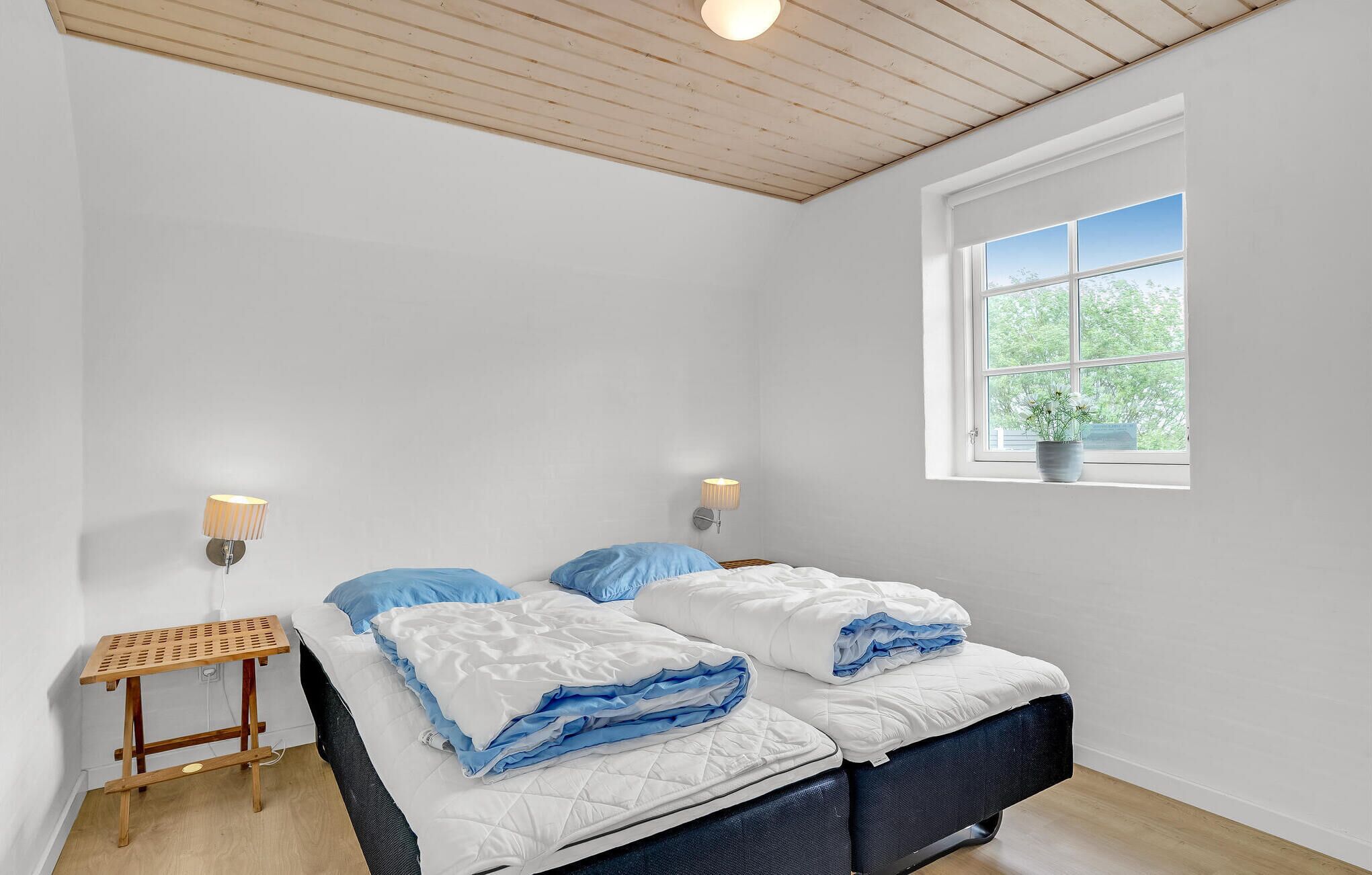 A bedroom with two beds, a window with a view of greenery, and a small table with a lamp.