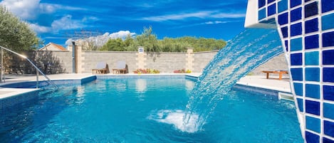 Piscina con una fuente de agua azul cristalina, dos sillas de descanso y un muro de piedra al fondo.