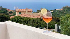 En balkong med ett glas vin och en citronskiva, med utsikt över ett kustlandskap med byggnader som har tak av terrakotta och frodig grönska.