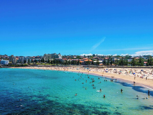 A beach with clear turquoise water, a sandy shore, and buildings in the background.