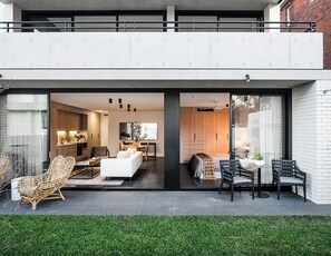 A modern house with a balcony, a living room with a sofa and chairs, a dining area with a table and chairs, and a kitchen visible through glass doors.