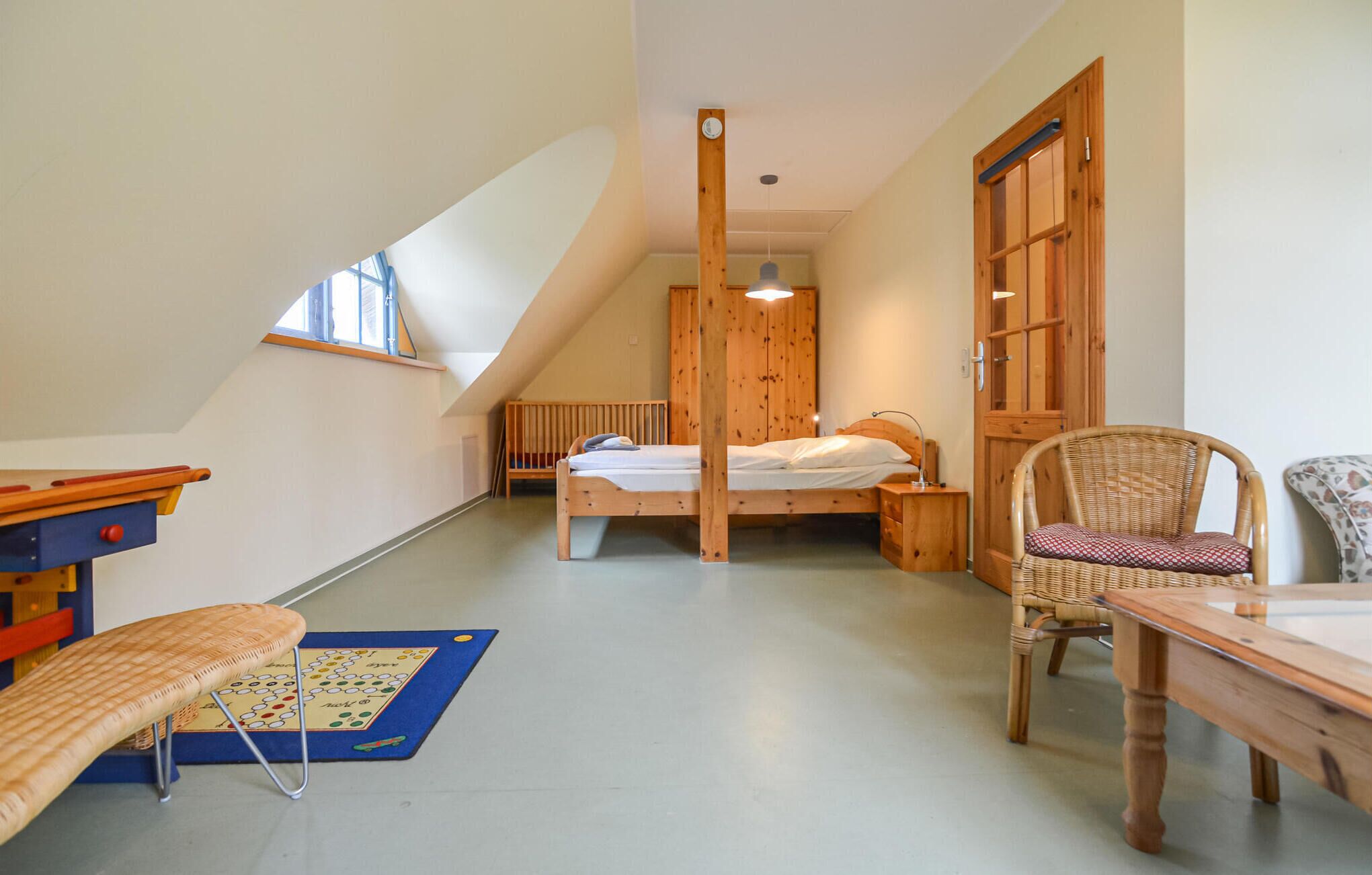 Een gezellig slaapkamer op zolder met een schuine kap, een houten bed, een klein tafeltje, een rieten stoel en een raam dat voor natuurlijk licht zorgt.