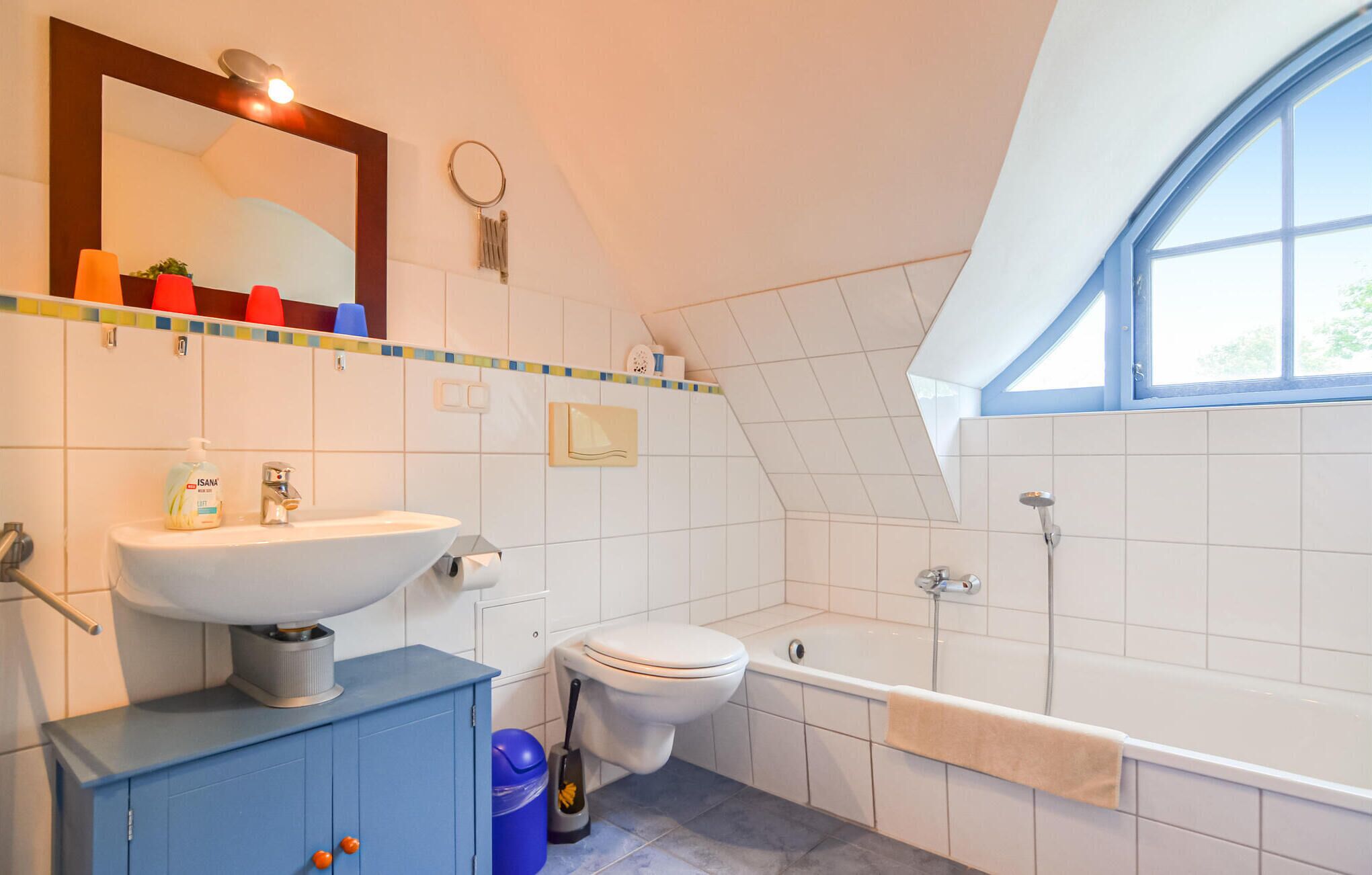 Une salle de bain avec un lavabo, des toilettes et une baignoire, tous de couleur blanche. Il y a une fenêtre donnant sur des arbres, un miroir au-dessus du lavabo et un meuble bleu contenant une poubelle.