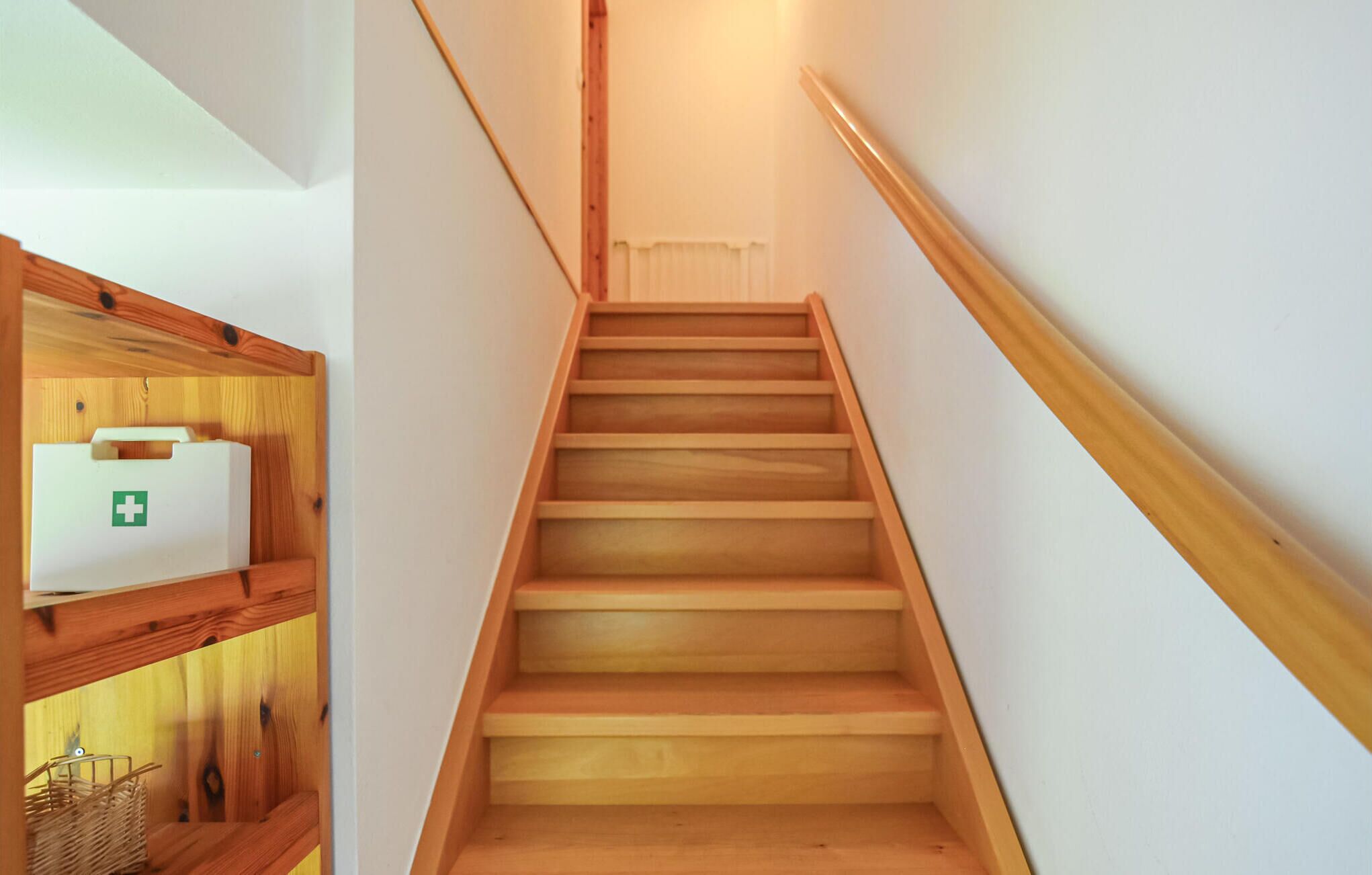 A wooden staircase with a handrail, leading to a landing with a white wall and a white object with a green cross on it.
