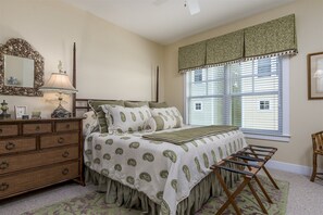 Ein Schlafzimmer mit Bett, Kommode, Spiegel, Lampe, Fenster mit Jalousien und einem Sessel.
