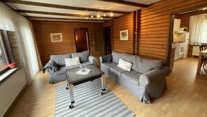 A cozy log cabin interior with two grey sofas, a small coffee table, and a kitchen area visible through an open doorway.