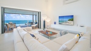 A modern living room with a large sofa, a coffee table, and a TV. There is a view of the ocean through a glass door.