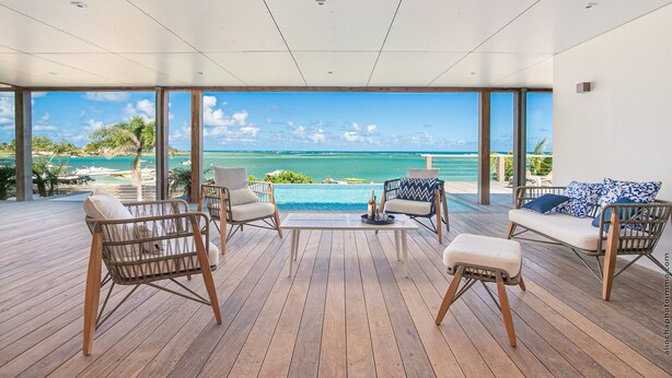 A modern outdoor lounge area with wicker furniture and a wooden deck, overlooking a clear blue sea and a tropical landscape.