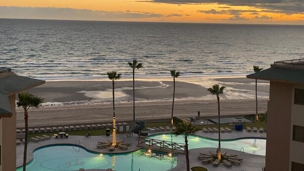 Ein Resort mit Poolanlage, Palmen und Blick auf den Ozean bei Sonnenuntergang.