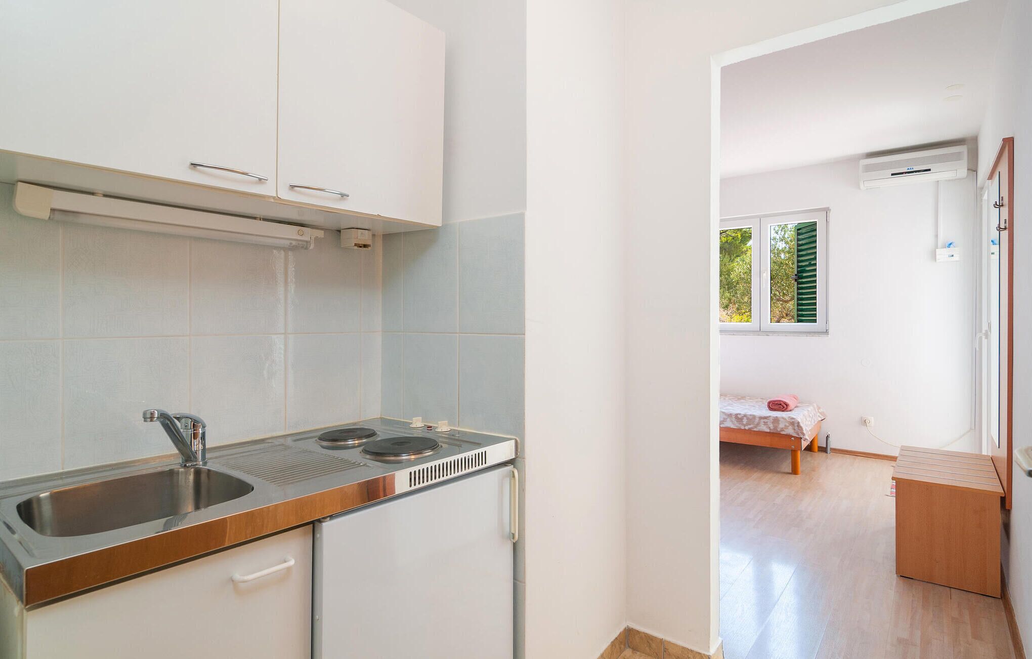 Una cocina compacta con un lavamanos, estufa y armarios. Se ve una cama pequeña con ventana y un mueble de madera en la habitación contigua.