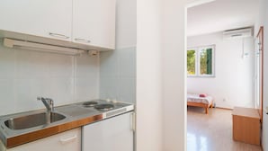 Una cocina compacta con un lavamanos, estufa y armarios. Se ve una cama pequeña con ventana y un mueble de madera en la habitación contigua.