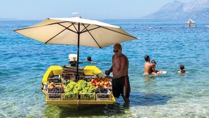 Un vendeur propose des fruits sous un parasol à bord d’un bateau, sur des eaux d’un bleu limpide, tandis que des gens nagent et que d’autres bateaux sont visibles au loin.
