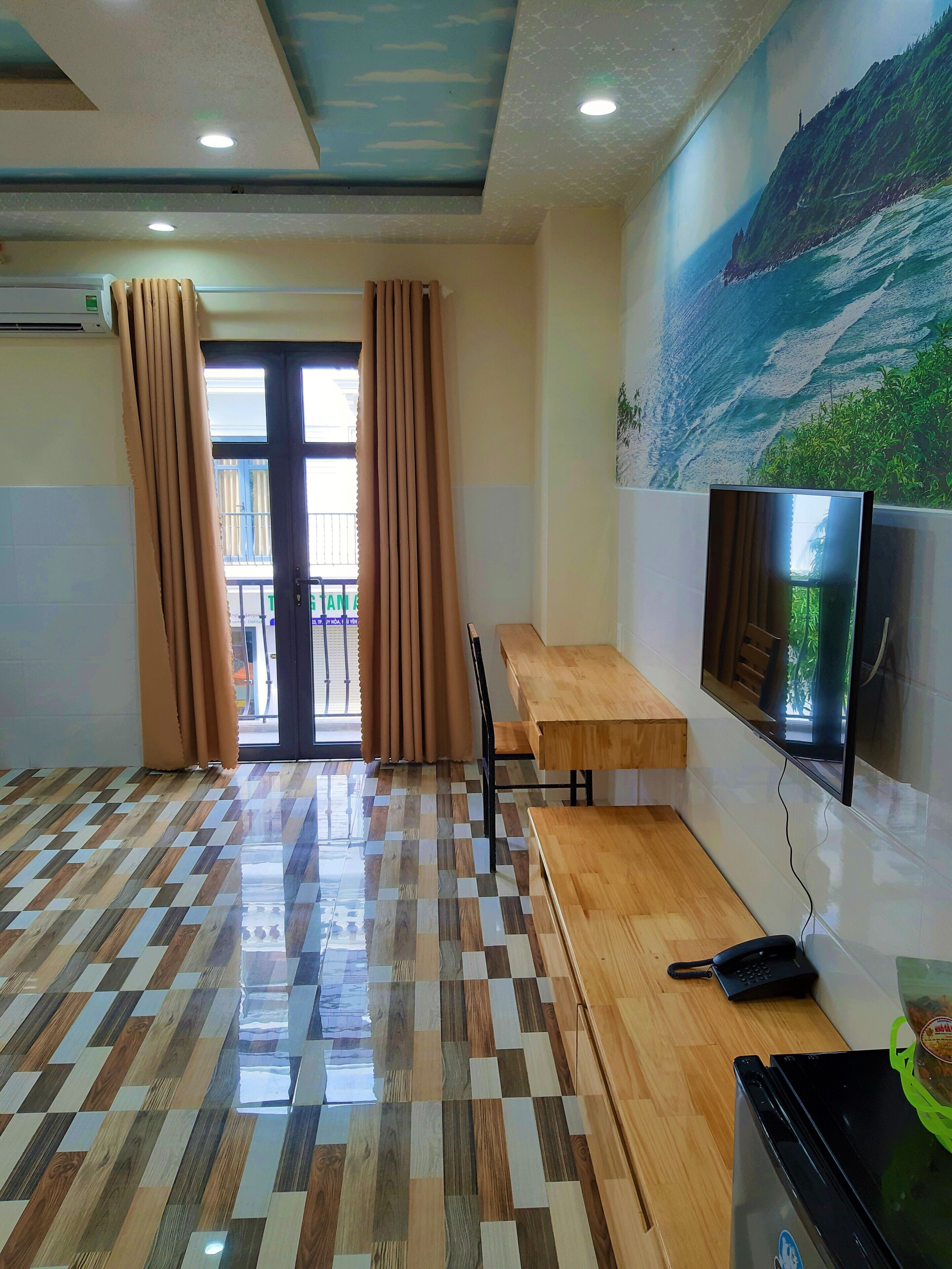 A room with a checkered wooden floor, a television mounted on the wall, a desk, and a chair.